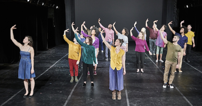 A group of dancers stand en mass in different colored-street clothes. They each outstretch their right arm. Their fingers angle toward them.